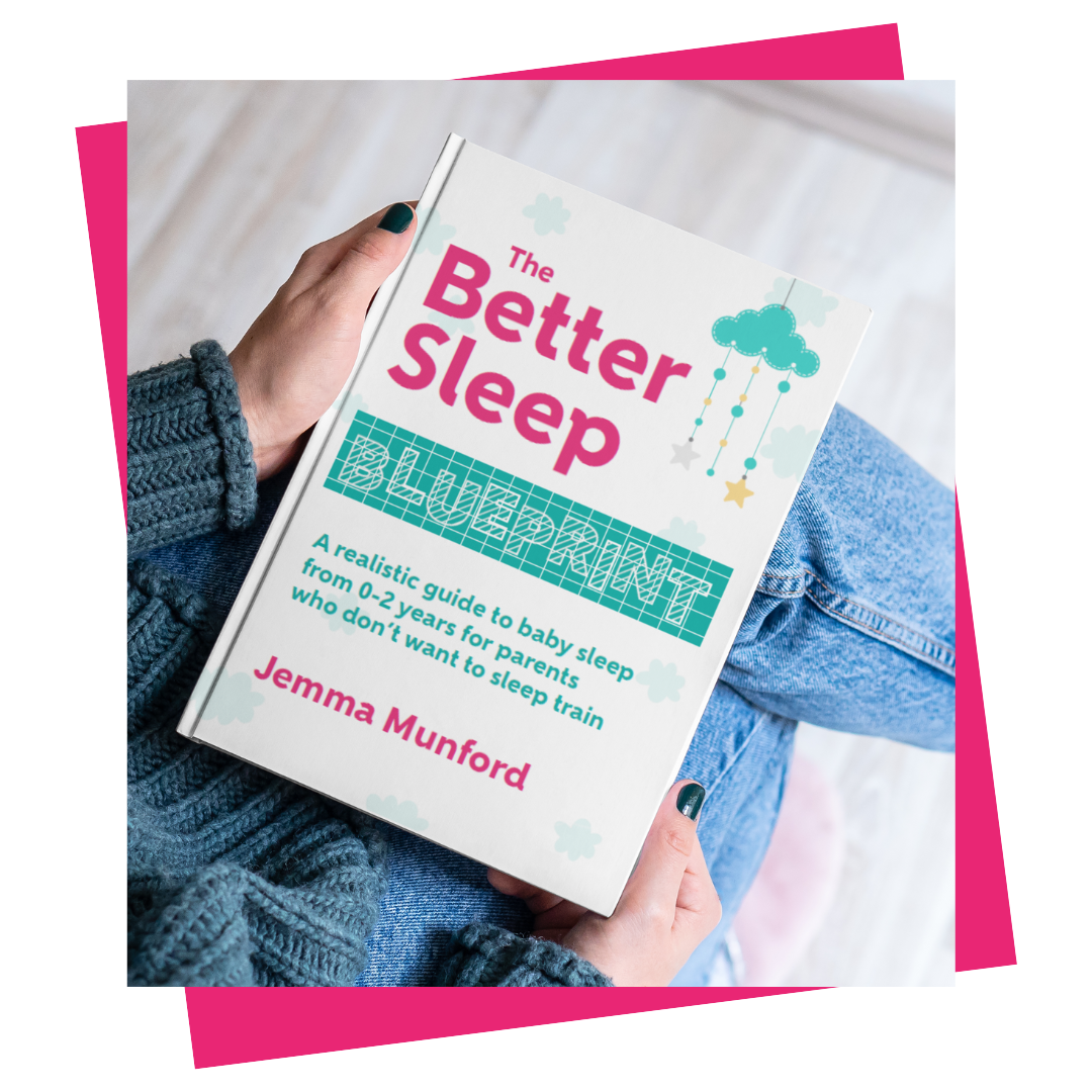 Female hands holding a book resting on a knee. The book is The Better Sleep Blueprint: A realistic guide to baby sleep for parents who don't want to sleep train. The cover is white with pink and teal writing.